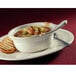 A Libbey cream white porcelain casserole dish with grilled bread on a plate with a spoon and a bowl of soup.