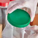 A hand in a glove holding a green 110/400 flat top lid over a plastic container.