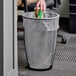 A hand putting a coffee cup in a Safco black round mesh wastebasket.