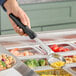 A hand holding a black Vollrath Spoodle over a tray of food.