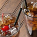 A close-up of a Eco-Products Black Unwrapped Compostable Plastic Straw in a glass with ice and brown liquid with a cherry on top.