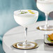 Two Acopa Deco coupe cocktail glasses on a table with white cocktails and herb garnishes.