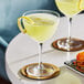 Two Acopa Deco Nick and Nora cocktail glasses with yellow liquid and lemon wedges on a white table.