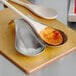 A Tablecraft brushed stainless steel double spoon rest holding a metal spoon and a wooden spoon with red sauce.