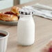 An Acopa glass milk bottle with a lid on a table next to a cup of coffee.