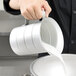 A hand using a Vollrath aluminum measuring cup to pour milk into a white bowl.