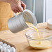 A hand pouring liquid from a Vollrath aluminum measuring cup into a bowl.