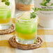 A glass of green and yellow liquid with a slice of apple on top with a bottle of Real Apple Puree Infused Syrup.