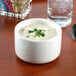 A Tuxton eggshell china soup cup filled with soup and garnished with parsley on a white background next to a glass of water.