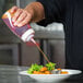 A hand using a Tablecraft Clear WideMouth Squeeze Bottle to pour sauce onto a plate of food.