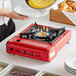 A person cooking food on a red Choice butane portable stove with a flame on top.