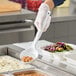 A person in gloves holding a white Vollrath Spoodle over food in a white container.
