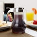 A Tablecraft syrup bottle with a chrome metal lid on a table next to a plate of food.