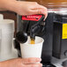 A hand using an Avantco faucet to pour coffee into a white cup.