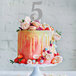 A cake with flowers and a silver glitter number 5 on top.