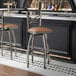 A metal bar stool with a cross back design and a light brown vinyl padded seat, featuring a swivel function.