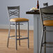 A Lancaster Table & Seating clear coat finish cross back bar stool with a light brown vinyl padded seat next to a table.