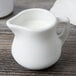 A Tuxton white porcelain creamer with a handle on a wood table filled with milk.