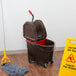 A Rubbermaid brown mop bucket with a red dirty water bucket and metal wringer on a wood floor.