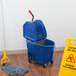 A Rubbermaid blue mop bucket with a handle next to a mop on a wood floor.
