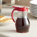 A clear polycarbonate teardrop pitcher of syrup with a red top.