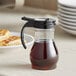 A Vollrath clear polycarbonate teardrop syrup server with black top filled with brown syrup on a table with food.