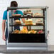 A man standing behind a Vollrath glass countertop display case with cakes.
