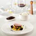 An ivory RAK Porcelain deep plate with meat and rice on it on a table with wine glasses.