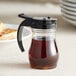 A clear polycarbonate teardrop syrup server with a black top and brown syrup inside on a table.