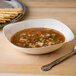 A Fineline ivory plastic bowl filled with soup next to a spoon and a plate of crackers.