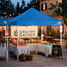 A blue 10' x 10' straight leg pop-up canopy tent set up at an outdoor market.