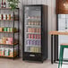 A black refrigerated glass door merchandiser with a left hinged door and LED lighting, stocked with various beverages.