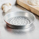 An American Metalcraft Super Perforated Pizza Pan on a counter next to a wooden cutting board and garlic bulbs.