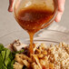 A hand pouring Ken's Foods Asian Sesame Dressing over a bowl of food.