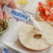 A hand holding a Philadelphia Original Plain Cream Cheese portion packet being spread over a bagel.