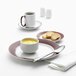 A white mug with a brown handle on a table with a bowl of soup and a spoon.