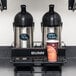 A Bunn Universal Airpot Rack holding two metal containers with black lids on a black shelf.