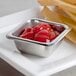 A square stainless steel sauce cup filled with ketchup on a table with a container of french fries.