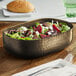 An American Metalcraft black hammered aluminum oval bowl filled with salad on a table.