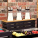 A GET metal gray beverage display crate holding three glass containers of white liquid on a hotel buffet table.