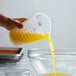 A hand using a Tablecraft flexible measuring cup to pour yellow liquid into a bowl.