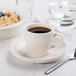 A CAC ivory scalloped edge espresso cup on a saucer with a spoon and a cup of coffee.