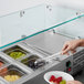 A hand holding a bowl of fruit over a refrigerated countertop prep rail.