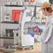 A man in a white coat and blue gloves using a Backyard Pro countertop vertical band meat saw to cut meat.
