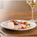A RAK Porcelain sand white flat coupe plate on a table with food and a glass of wine.