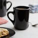 A black mug with a handle on a white surface with a plate of food and a black mug with a handle and a spoon.