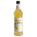 A bottle of Monin Key Lime Pie flavoring syrup in front of a glass of yellow liquid with mint on a white background.