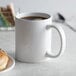 A close-up of a white Acopa stoneware mug filled with brown liquid next to a donut.