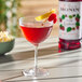 A glass of red raspberry-flavored drink garnished with a lemon twist and raspberry, with a bottle of Monin Premium Wild Raspberry Flavoring Syrup in the background.