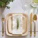 A table setting with a Bambu square bamboo plate, gold cutlery, and a napkin with a sprig of greenery tied to it.